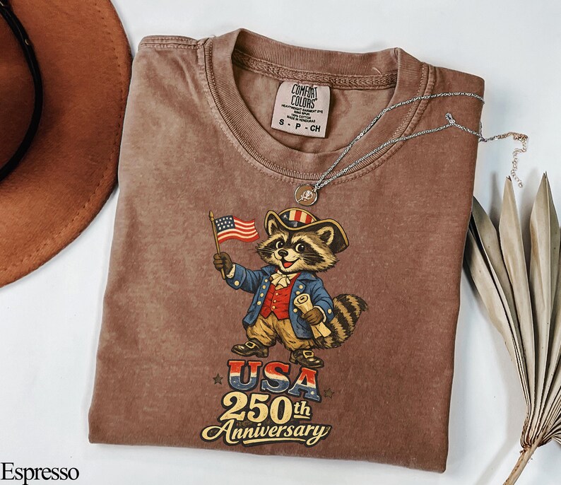 Comfort Colors 1717 garment-dyed t-shirt in Espresso brown featuring a vintage-style 'USA 250th Anniversary' graphic. The illustration depicts a patriotic raccoon 'trash panda' dressed as a Founding Father in Revolutionary War attire holding an American flag. Perfect retro Semiquincentennial gift for history lovers celebrating America's 250th birthday.