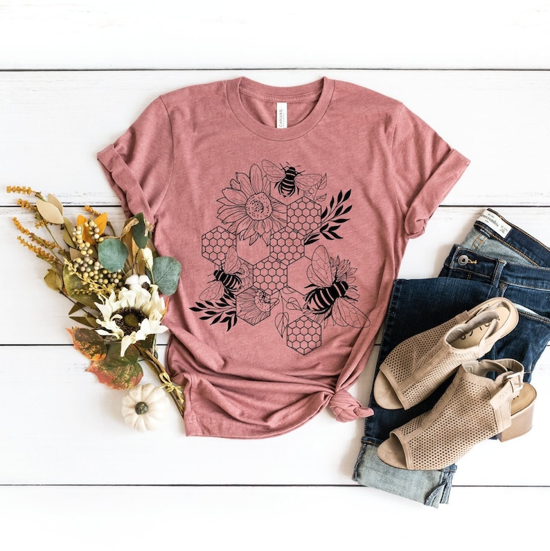 May include: A mauve t-shirt with a black and white graphic design of bees and honeycombs. The design features a sunflower and leaves.