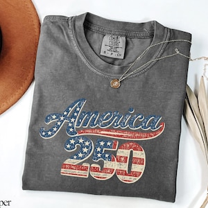 Pode incluir: Uma camiseta cinza Comfort Colors com a palavra "America" em estrelas azuis e brancas e listras vermelhas, e o n&uacute;mero "250" em listras vermelhas e brancas. Um colar de ouro com um pingente est&aacute; na camisa. Um chap&eacute;u marrom e folhas de palmeira secas est&atilde;o no fundo.