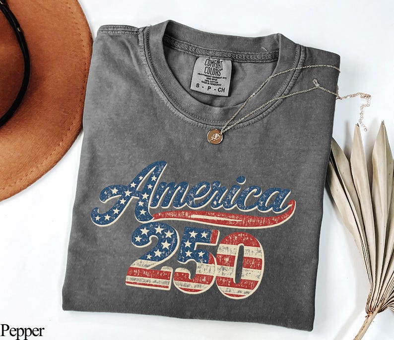 Pode incluir: Uma camiseta cinza Comfort Colors com a palavra "America" em estrelas azuis e brancas e listras vermelhas, e o n&uacute;mero "250" em listras vermelhas e brancas. Um colar de ouro com um pingente est&aacute; na camisa. Um chap&eacute;u marrom e folhas de palmeira secas est&atilde;o no fundo.