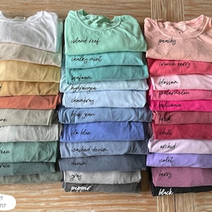 Pode incluir: Pilhas de camisetas Comfort Colors 1717 em v&aacute;rias cores, incluindo branco, mostarda e azul jeans. Cada camisa &eacute; rotulada com o nome de sua cor em um estilo manuscrito. As camisas s&atilde;o cuidadosamente dobradas e empilhadas em uma superf&iacute;cie de madeira.
