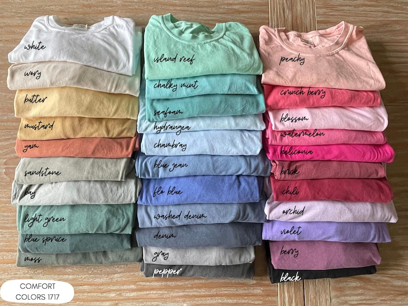 Pode incluir: Pilhas de camisetas Comfort Colors 1717 em v&aacute;rias cores, incluindo branco, mostarda e azul jeans. Cada camisa &eacute; rotulada com o nome de sua cor em um estilo manuscrito. As camisas s&atilde;o cuidadosamente dobradas e empilhadas em uma superf&iacute;cie de madeira.