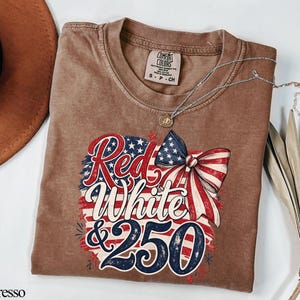 May include: Espresso-colored t-shirt with a patriotic design featuring the text "Red, White & 250" in red and blue, with a bow and stars. A silver necklace with a pendant is on the shirt.