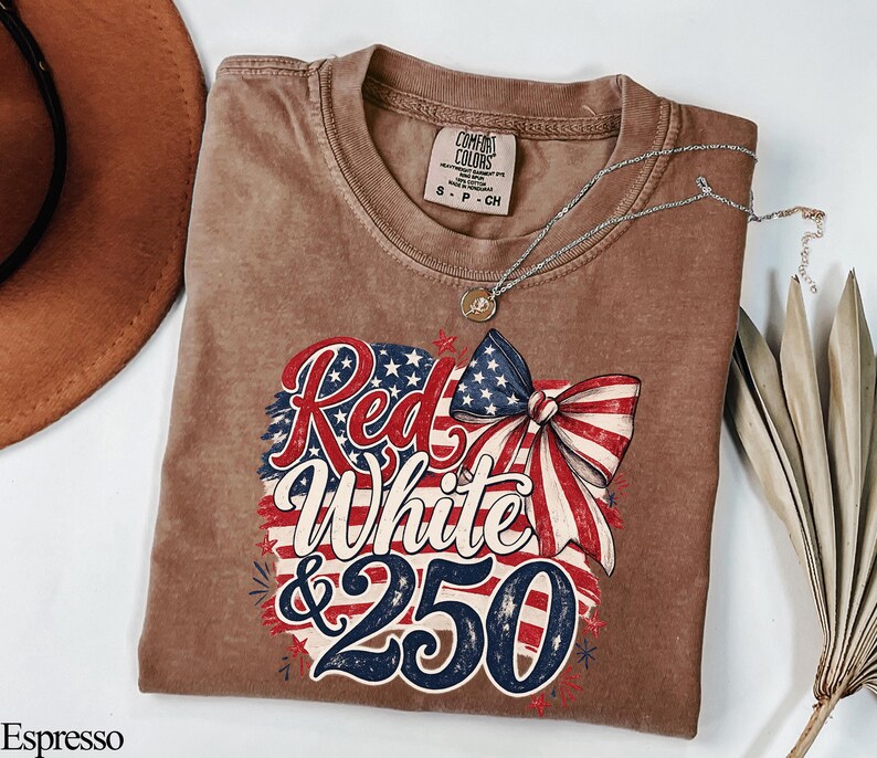 May include: Espresso-colored t-shirt with a patriotic design featuring the text "Red, White & 250" in red and blue, with a bow and stars. A silver necklace with a pendant is on the shirt.