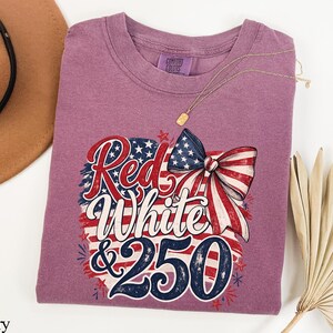 May include: Berry-colored t-shirt with a patriotic design featuring a bow and the words "Red, White & 250". The design incorporates the American flag's colors and stars. A gold necklace is visible.