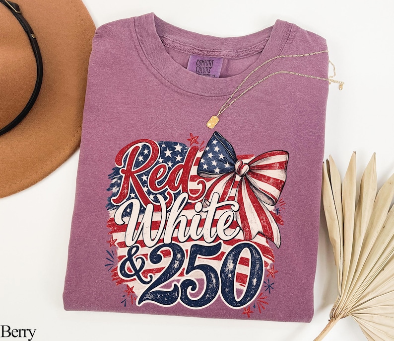 May include: Berry-colored t-shirt with a patriotic design featuring a bow and the words "Red, White & 250". The design incorporates the American flag's colors and stars. A gold necklace is visible.