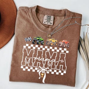 May include: Brown t-shirt with the word "MAMA" in white block letters over a checkered pattern. The shirt features illustrations of monster trucks in green, yellow, and red, and a checkered flag. A silver necklace is draped across the shirt.