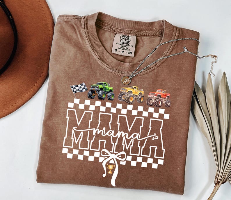 May include: Brown t-shirt with the word "MAMA" in white block letters over a checkered pattern. The shirt features illustrations of monster trucks in green, yellow, and red, and a checkered flag. A silver necklace is draped across the shirt.
