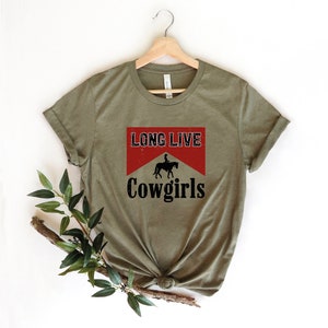May include: Olive green t-shirt with the text "LONG LIVE Cowgirls" in a red and white design. The design includes a silhouette of a person riding a horse. The shirt is displayed on a wooden hanger.