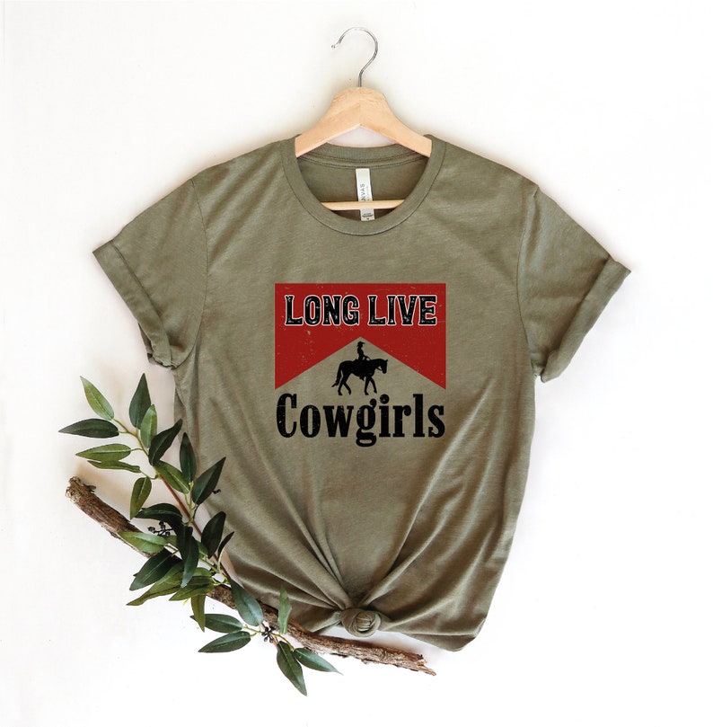 May include: Olive green t-shirt with the text "LONG LIVE Cowgirls" in a red and white design. The design includes a silhouette of a person riding a horse. The shirt is displayed on a wooden hanger.