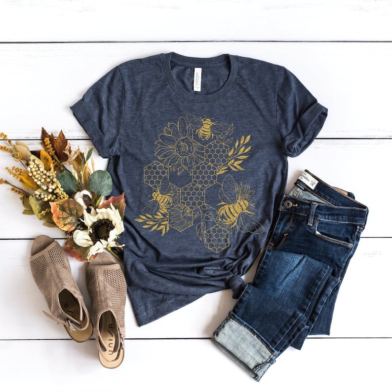 May include: A navy blue t-shirt with a gold honeycomb design featuring bees and sunflowers. The shirt is laid flat on a white wooden surface with a pair of brown sandals and blue jeans.
