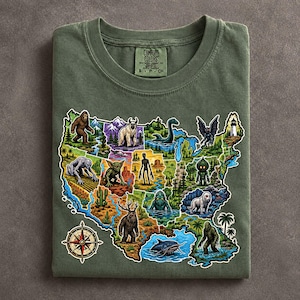 May include: An olive green t-shirt featuring a graphic map of the United States populated with various cryptids and mythical creatures. The design includes a compass rose in the bottom left corner and a Comfort Colors brand label at the neck.
