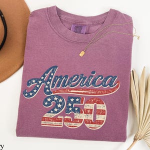Pode incluir: T-shirt cor de baga com o texto "America 250" num design patri&oacute;tico. O texto apresenta um esquema de cores vermelho e azul com estrelas e listras, evocando uma est&eacute;tica vintage. Um colar de ouro est&aacute; drapeado sobre a camisa.