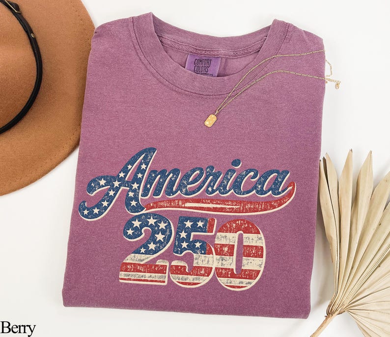 Pode incluir: T-shirt cor de baga com o texto "America 250" num design patri&oacute;tico. O texto apresenta um esquema de cores vermelho e azul com estrelas e listras, evocando uma est&eacute;tica vintage. Um colar de ouro est&aacute; drapeado sobre a camisa.