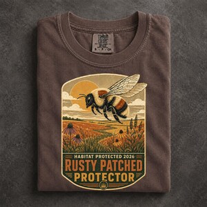May include: A brown t-shirt featuring a graphic of a bumblebee flying over a field with a sun in the background. The text reads HABITAT PROTECTED 2026 RUSTY PATCHED PROTECTOR. The shirt has a Comfort Colors brand label at the inner neckline.