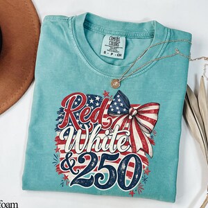 May include: Seafoam green t-shirt with a patriotic design. The graphic features the words "Red White & 250" in red, white, and blue, along with a bow and stars. A gold necklace is draped across the shirt.