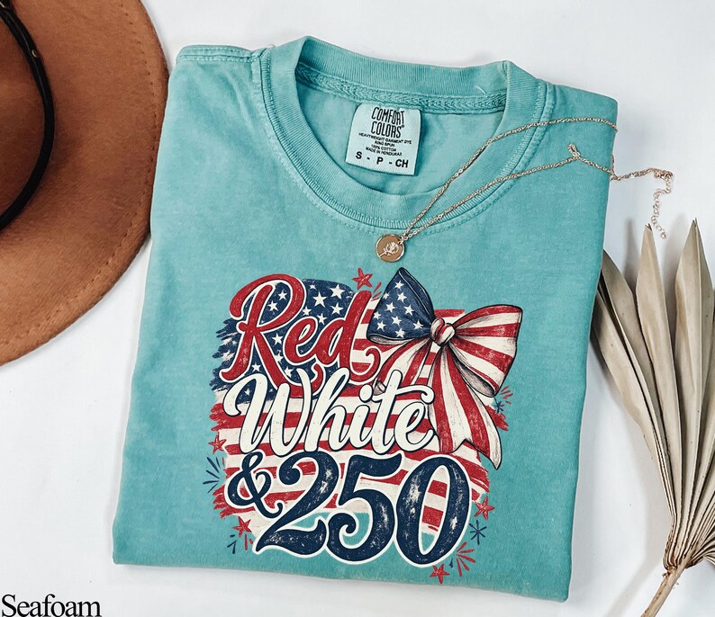 May include: Seafoam green t-shirt with a patriotic design. The graphic features the words "Red White & 250" in red, white, and blue, along with a bow and stars. A gold necklace is draped across the shirt.