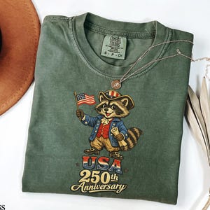 A Comfort Colors 1717 garment-dyed tee in Moss green featuring a vintage-style raccoon dressed in a Revolutionary War colonial uniform holding an American flag. The graphic displays 'USA 250th Anniversary' in retro typography, ideal for the 2026 Semiquincentennial and patriotic history enthusiasts.