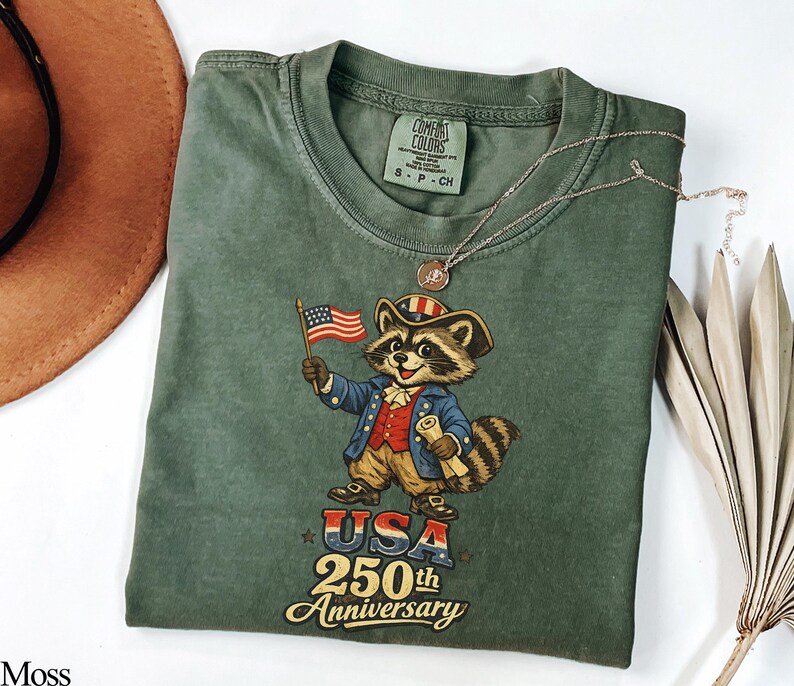 A Comfort Colors 1717 garment-dyed tee in Moss green featuring a vintage-style raccoon dressed in a Revolutionary War colonial uniform holding an American flag. The graphic displays 'USA 250th Anniversary' in retro typography, ideal for the 2026 Semiquincentennial and patriotic history enthusiasts.