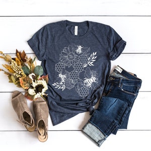 May include: A navy blue t-shirt with a white graphic design of a honeycomb, bees, and sunflowers. The shirt is laid flat on a white wooden surface with a pair of brown sandals and blue jeans.