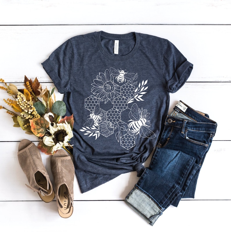 May include: A navy blue t-shirt with a white graphic design of a honeycomb, bees, and sunflowers. The shirt is laid flat on a white wooden surface with a pair of brown sandals and blue jeans.