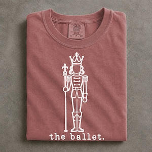 May include: A dusty rose crew neck t-shirt featuring a white line art graphic of a nutcracker holding a staff. The text "the ballet." is printed below the nutcracker. The inner neck label reads "Comfort Colors" with size S-CH.