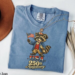 Comfort Colors 1717 garment-dyed t-shirt in Blue Jean featuring a vintage Colonial Raccoon dressed as a Founding Father holding an American flag. The graphic includes the text 'USA 250th Anniversary' in a retro patriotic font. This Semiquincentennial shirt is perfect for history buffs and 1776-2026 Independence Day celebrations.