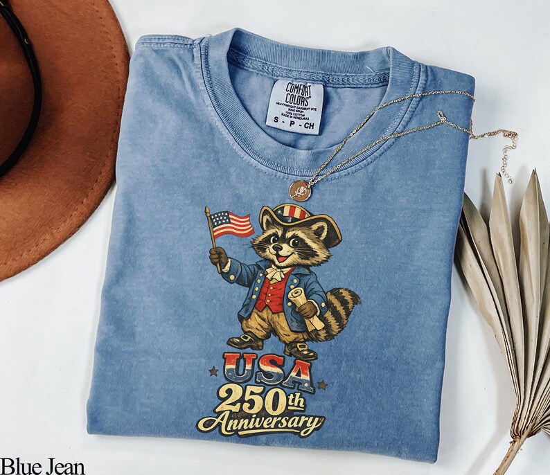 Comfort Colors 1717 garment-dyed t-shirt in Blue Jean featuring a vintage Colonial Raccoon dressed as a Founding Father holding an American flag. The graphic includes the text 'USA 250th Anniversary' in a retro patriotic font. This Semiquincentennial shirt is perfect for history buffs and 1776-2026 Independence Day celebrations.