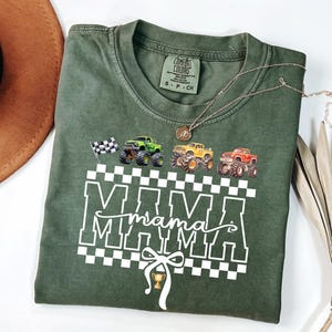 May include: Green Comfort Colors t-shirt with a graphic of monster trucks and the word "MAMA" in white. The design includes a checkered pattern, a checkered flag, and a trophy. A gold necklace is draped across the shirt.