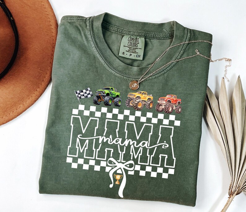 May include: Green Comfort Colors t-shirt with a graphic of monster trucks and the word "MAMA" in white. The design includes a checkered pattern, a checkered flag, and a trophy. A gold necklace is draped across the shirt.
