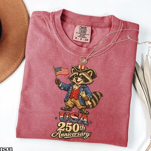Comfort Colors 1717 t-shirt in Crimson featuring a vintage-style colonial raccoon in Revolutionary War attire holding an American flag and scroll. The graphic prominently displays 'USA 250th Anniversary' in retro typography. This patriotic trash panda apparel is the perfect Semiquincentennial gift for the 2026 independence celebration.