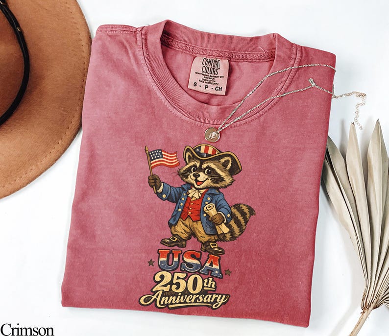Comfort Colors 1717 t-shirt in Crimson featuring a vintage-style colonial raccoon in Revolutionary War attire holding an American flag and scroll. The graphic prominently displays 'USA 250th Anniversary' in retro typography. This patriotic trash panda apparel is the perfect Semiquincentennial gift for the 2026 independence celebration.