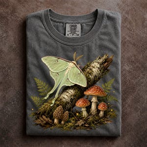 May include: A charcoal gray t-shirt featuring a graphic print of a pale green luna moth resting on a mossy birch log. The scene includes red-capped mushrooms, brown morel mushrooms, and green fern fronds. The inner neck label reads Comfort Colors.