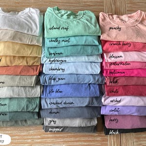 Pode incluir: Pilhas de camisetas dobradas em v&aacute;rias cores, incluindo branco, amarelo, verde, azul, rosa e roxo. Cada camisa &eacute; rotulada com o nome da cor em escrita preta. O texto "COMFORT COLORS 1717" est&aacute; na parte inferior.