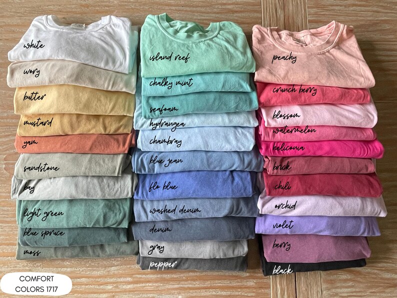 Pode incluir: Pilhas de camisetas dobradas em v&aacute;rias cores, incluindo branco, amarelo, verde, azul, rosa e roxo. Cada camisa &eacute; rotulada com o nome da cor em escrita preta. O texto "COMFORT COLORS 1717" est&aacute; na parte inferior.
