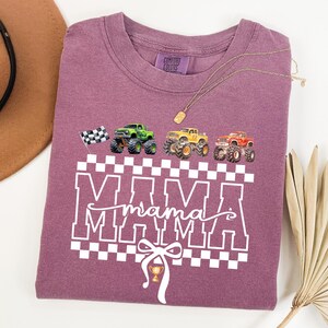 May include: A mauve-colored t-shirt with a graphic design featuring the word "MAMA" in white block letters, a checkered pattern, and illustrations of monster trucks. A checkered flag and a trophy are also included in the design.