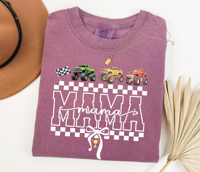 May include: A mauve-colored t-shirt with a graphic design featuring the word "MAMA" in white block letters, a checkered pattern, and illustrations of monster trucks. A checkered flag and a trophy are also included in the design.