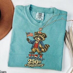 Comfort Colors 1717 Seafoam green shirt featuring a vintage-style illustration of a Founding Father raccoon holding a 1776 flag. Text reads 'USA 250th Anniversary' in a retro font. Ideal patriotic gift for the 2026 Semiquincentennial and history-loving trash panda fans.