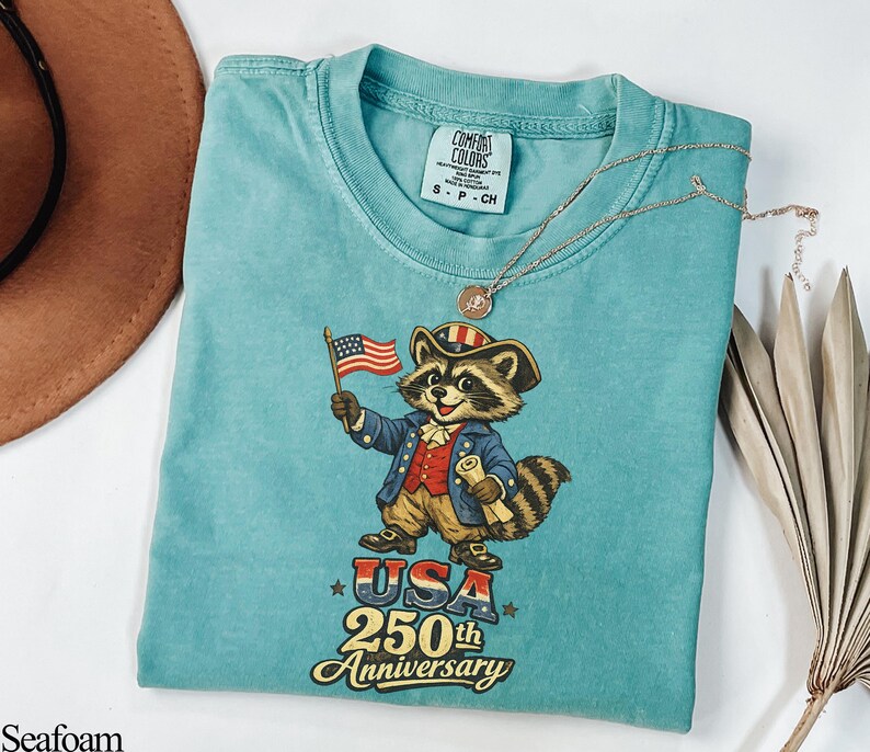 Comfort Colors 1717 Seafoam green shirt featuring a vintage-style illustration of a Founding Father raccoon holding a 1776 flag. Text reads 'USA 250th Anniversary' in a retro font. Ideal patriotic gift for the 2026 Semiquincentennial and history-loving trash panda fans.