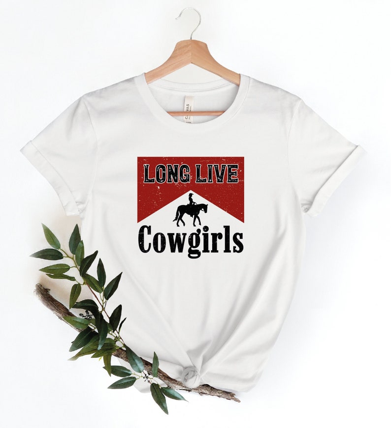 May include: White t-shirt with the text "LONG LIVE Cowgirls" in a red and black design. The design includes a silhouette of a person riding a horse. The shirt is hanging on a wooden hanger.
