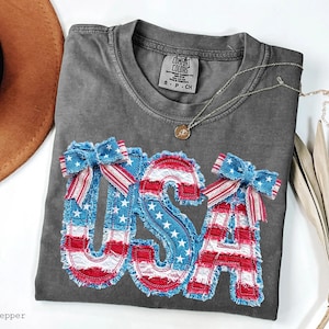 May include: A gray Comfort Colors t-shirt with a patriotic "USA" design in red, white, and blue. The letters are embellished with stars and bows. A gold necklace and a brown hat are also visible.