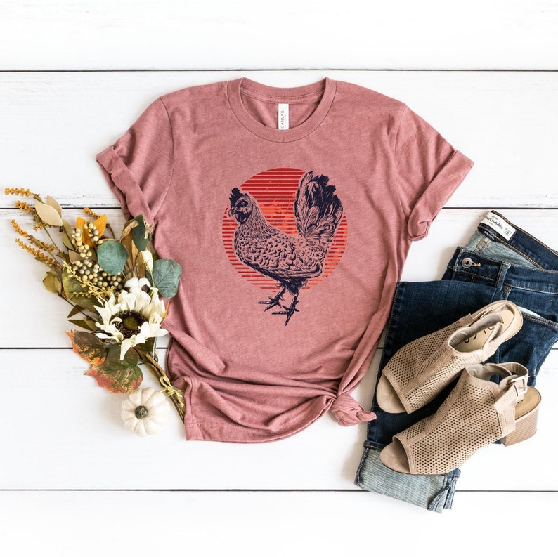 Puede incluir: Una camiseta color malva con un gr&aacute;fico de un gallo frente a un sol rojo. El gallo es blanco y negro con un contorno azul. La camiseta est&aacute; colocada sobre una superficie de madera blanca con un par de sandalias marrones y unos vaqueros azules.