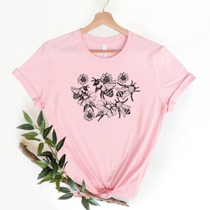 May include: A light pink t-shirt featuring a black and white illustration of bees and flowers. The graphic is centered on the front of the shirt. The shirt is hanging on a wooden hanger.
