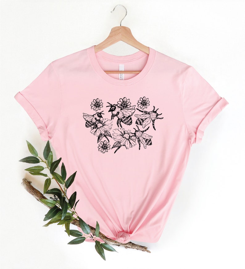 May include: A light pink t-shirt featuring a black and white illustration of bees and flowers. The graphic is centered on the front of the shirt. The shirt is hanging on a wooden hanger.