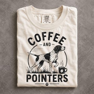 May include: An off-white crew neck t-shirt featuring a black graphic print of a spotted pointer dog standing in grass next to a steaming coffee mug. The text reads COFFEE AND POINTERS with a small paw print icon centered at the bottom of the design.
