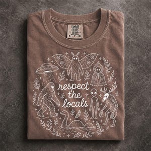 May include: A brown crew neck t-shirt featuring a white graphic print of various cryptids, including a mothman, bigfoot, alien, and a sea creature, surrounding the text "respect the locals" with small stars and botanical leaf accents.