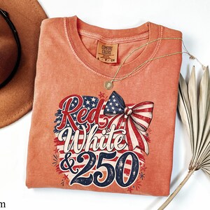 May include: A coral-colored t-shirt with a patriotic design. The graphic features the words "Red White & 250" in red, white, and blue, along with a bow and stars. A gold necklace and a brown hat are also visible.
