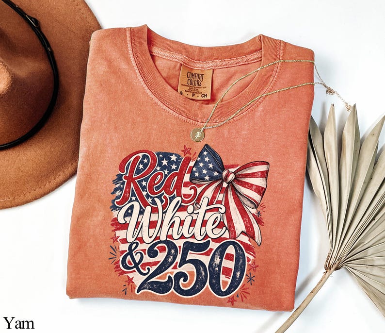 May include: A coral-colored t-shirt with a patriotic design. The graphic features the words "Red White & 250" in red, white, and blue, along with a bow and stars. A gold necklace and a brown hat are also visible.