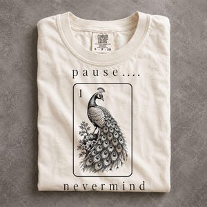 May include: A cream-colored short-sleeve t-shirt featuring a black graphic of a peacock inside a rectangular frame. Above the frame is the text "p a u s e . . . ." and below is the text "n e v e r m i n d". The number "1" is printed in the top left corner.