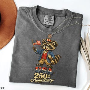 Comfort Colors 1717 t-shirt in Pepper grey featuring a vintage-style patriotic raccoon dressed as a Founding Father for the USA 250th Anniversary. The graphic shows a colonial 'Trash Panda' in a Revolutionary War uniform holding an American flag with text reading 'USA 250th Anniversary'. Perfect retro semiquincentennial gift for history buffs and Independence Day 2026 celebrations.